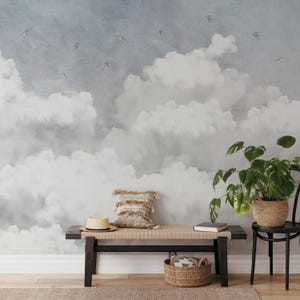 May include: A mural depicting a cloudy sky with birds in flight. A bench with a hat, pillow, and basket is in front of the mural. A potted plant in a woven basket sits on a black chair. The color palette is soft, with shades of gray, white, and blue.