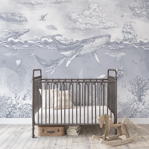 May include: A dark brown metal crib with a white mattress and a beige checkered pillow, positioned in front of a wall mural featuring gray-toned illustrations of whales, a sailing ship, coral reefs, and sea creatures under a cloudy sky.