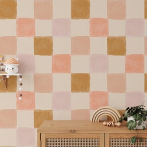 May include: A beige wall with a checkered pattern wallpaper. The wallpaper features large squares in shades of pink, peach, and brown. A wooden dresser with two drawers sits in front of the wall. A rainbow wooden toy and a potted plant are on top of the dresser.