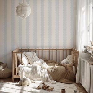 May include: A light wood crib with bedding, pillows, and a blanket. The room has a patterned wallpaper with vertical light blue scalloped stripes. A white paper lantern hangs from the ceiling. A rug and wooden toys are on the floor.