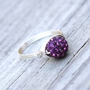 May include: A silver ring featuring a cluster of small, sparkling purple crystals. The ring has a simple, open band design, with the crystal cluster as the focal point, set in a wire-wrapped setting. The ring is displayed on a weathered, light-coloured wooden surface.