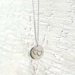 May include: A silver necklace with a circular pendant, engraved with two interlocking hearts. The pendant is suspended from a fine chain. The necklace is shown against a white, textured backdrop.