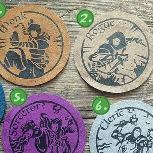 May include: Ten circular leather coasters in various colours, each featuring a black ink illustration of a fantasy character class and its name, including Monk, Rogue, Paladin, Wizard, Sorcerer, Cleric, Druid, Bard, and Warlock, labelled one through ten.