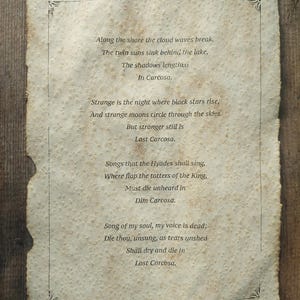 May include: A rectangular piece of aged, textured parchment paper with deckled edges, featuring a thin black border and the printed poem Cassilda's Song from The King in Yellow, beginning with the lines Along the shore the cloud waves break.