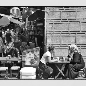 May include: Black and white street scene with people at tables and a shop in the background. Two women sit at a small table, engaged in conversation. A Turkish flag hangs above a shop with Coca-Cola signage.