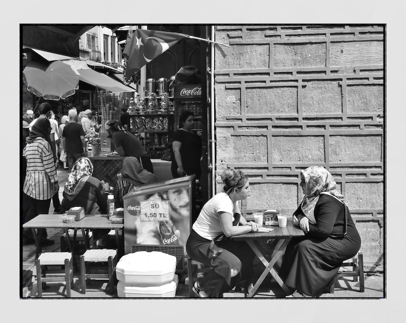 May include: Black and white street scene with people at tables and a shop in the background. Two women sit at a small table, engaged in conversation. A Turkish flag hangs above a shop with Coca-Cola signage.