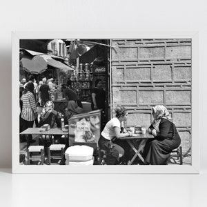 May include: Black and white photograph of a street scene with people. Two women sit at a small table, engaged in conversation. Other people are in the background near a shop with a Coca-Cola sign.