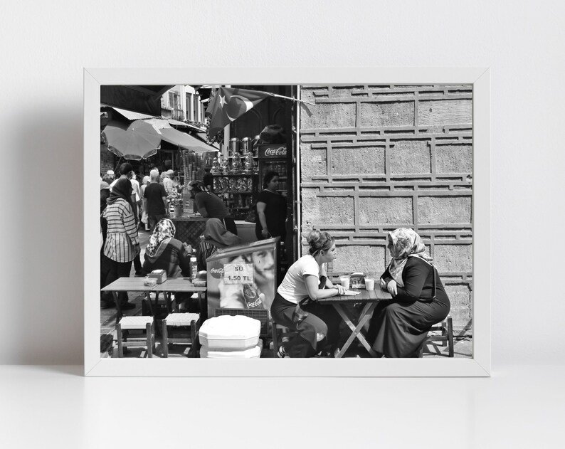 May include: Black and white photograph of a street scene with people. Two women sit at a small table, engaged in conversation. Other people are in the background near a shop with a Coca-Cola sign.