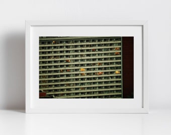 May include: A photograph of a large apartment building with many windows. Some of the windows are lit up, while others are dark. The building is made of concrete and has a simple design.