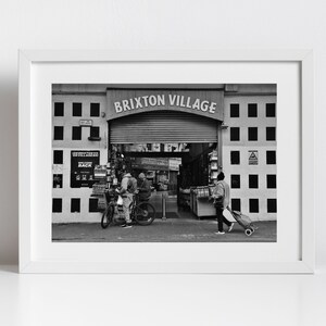 Könnte beinhalten: Schwarzweißfoto eines Markteingangs mit den Worten "BRIXTON VILLAGE" über einem Rolltor. Zwei Personen gehen durch den Eingang, eine schiebt einen Einkaufswagen.