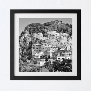 May include: Black and white framed print of a coastal town nestled on a hillside. The image features a cluster of buildings with a mountain backdrop, creating a scenic landscape. The print is encased in a black frame.
