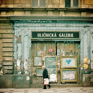 May include: A storefront with a sign that reads "Uličnick&aacute; Galerie" in a faded blue and white striped awning. The storefront is covered in peeling paint and graffiti. A man in a dark suit walks past the storefront.
