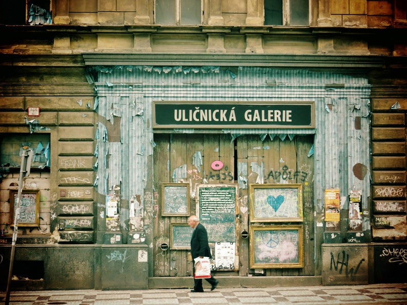May include: A storefront with a sign that reads "Uličnick&aacute; Galerie" in a faded blue and white striped awning. The storefront is covered in peeling paint and graffiti. A man in a dark suit walks past the storefront.