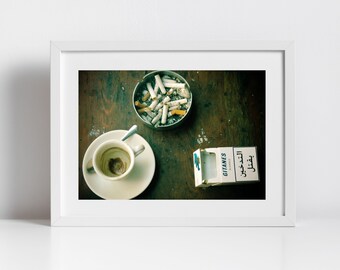 May include: A framed print featuring a still life with a used coffee cup on a saucer, a metal ashtray filled with cigarette butts, and a partially open pack of Gitanes Blondes cigarettes. The items are arranged on a dark wooden surface.