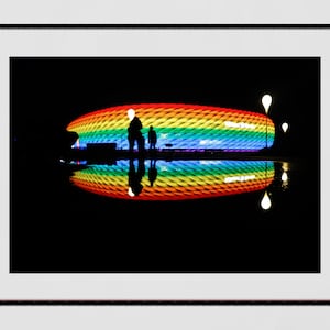 Peut inclure: Un grand stade illuminé d'un motif arc-en-ciel la nuit. Le stade se reflète dans une étendue d'eau devant lui.