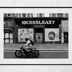 Könnte beinhalten: Ein Schwarzweißfoto eines Motorrads, das eine Straße entlangfährt, vor einem Gebäude mit dem Schriftzug "NICSLEAZY" an der Vorderseite. Das Gebäude hat Graffiti an der Seite, darunter ein Comic-Gesicht mit den Worten "LOVE MORE".