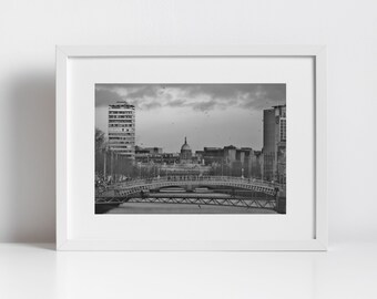 May include: A black and white photograph of a bridge over a river in Dublin, Ireland. The city skyline is in the background, including the iconic Dublin Spire.