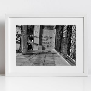 May include: Black and white photograph of a graffiti-covered alleyway with the words "The Revolution Must Be Feminist" written on a wall.