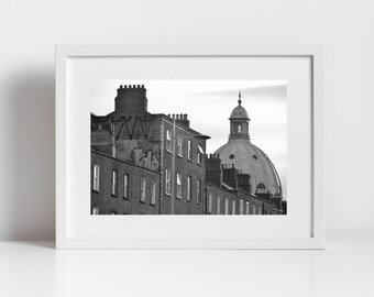 May include: Black and white photograph of a city street with a large domed building in the background. The building has a tall spire and is surrounded by smaller buildings.