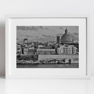 May include: A black and white photograph of a cityscape with a large domed building in the centre. The city is built on a peninsula with a body of water in the foreground.