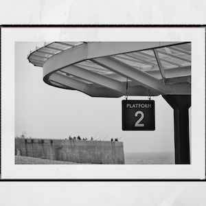 May include: Black and white photograph of a platform sign that reads "PLATFORM 2" hanging from a metal structure. The sign is hanging over a body of water with a concrete wall in the background.