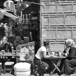 May include: Black and white street scene with people at tables, a Coca-Cola cooler, and a Turkish flag. Two women sit at a table, engaged in conversation. A shop with various items is visible in the background.