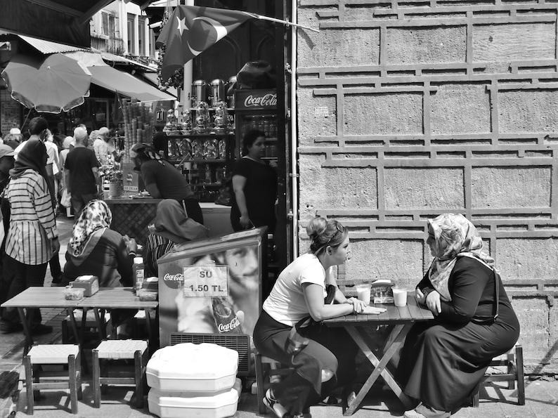 May include: Black and white street scene with people at tables, a Coca-Cola cooler, and a Turkish flag. Two women sit at a table, engaged in conversation. A shop with various items is visible in the background.