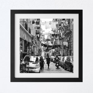 May include: A black and white photograph of a narrow street in a city. Clothes are hanging on lines strung across the street. Two people walk down the street, and a motorcycle is parked in the street.