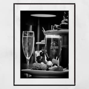 May include: A black and white photo of a glass of champagne and a glass of beer with the brand name "Moritz" on it. The glasses are on a plate with olives and other snacks.