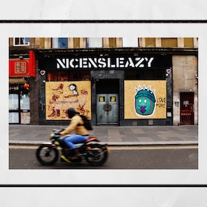 Könnte beinhalten: Straßenszene mit einem Gebäude mit dem Text "NICENSLIEAZY". Zwei Holztafeln zeigen Graffiti-Kunst. Ein Motorrad bewegt sich auf der Straße. Das Bild ist schwarz gerahmt und fängt einen Moment des Stadtlebens ein.