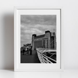 Puede incluir: Fotografía en blanco y negro de un gran edificio industrial con el texto "BALTIC FLOUR MILLS" en el lateral. Personas caminando por un puente en primer plano.