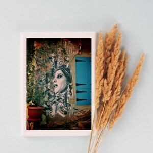 May include: A framed print of a colorful street art mural featuring a woman's face with a blue door in the background.