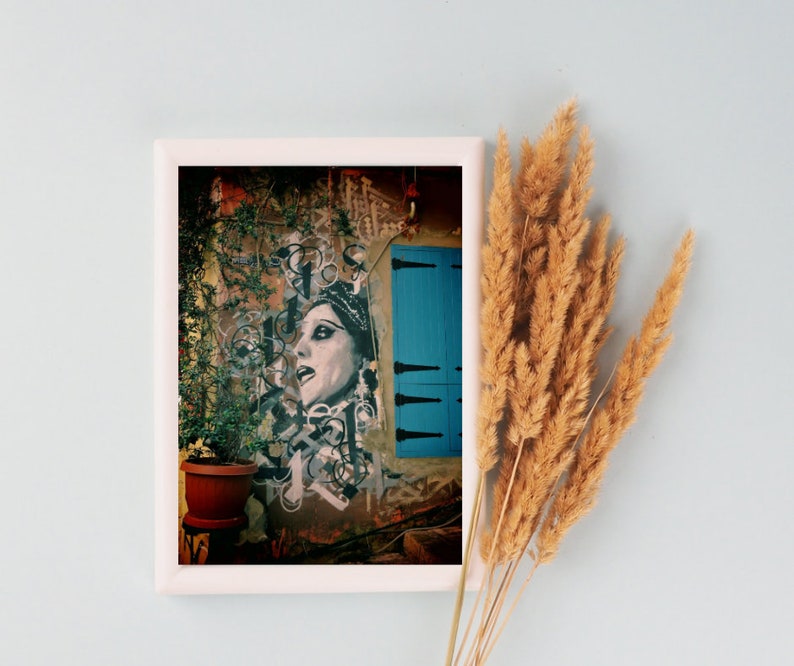 May include: A framed print of a colorful street art mural featuring a woman's face with a blue door in the background.