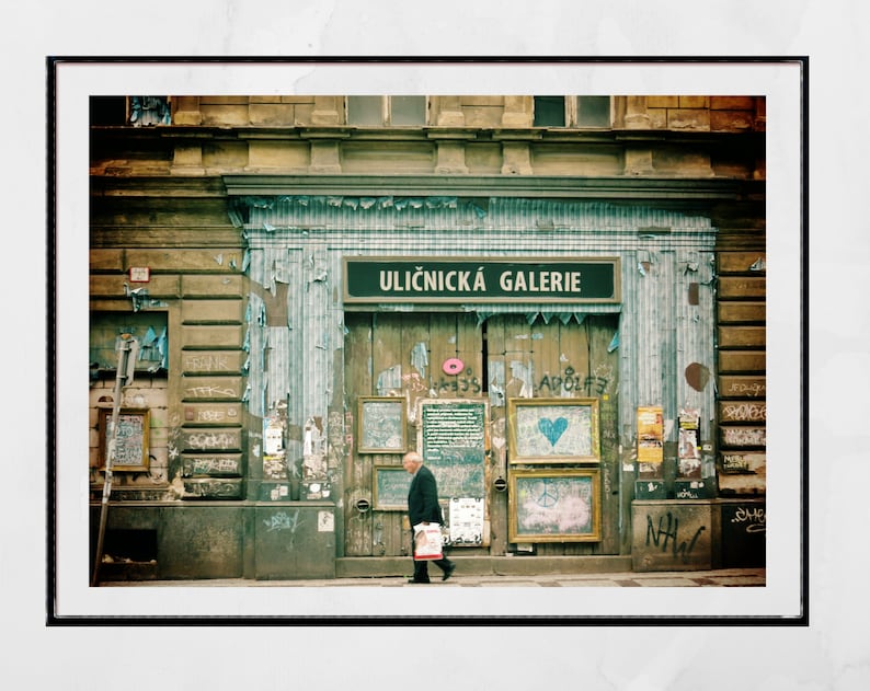 May include: A weathered building with a sign that reads "Uličnick&aacute; Galerie" in black letters on a white background. The building has a wooden door with several posters and signs on it. A person is walking in front of the building.
