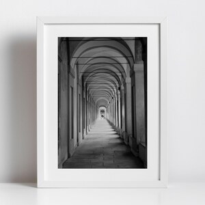Könnte beinhalten: Schwarz-weiß gerahmte Fotografie eines langen, gewölbten Korridors. Die Perspektive lenkt den Blick entlang des Korridors, wobei sich wiederholende Bögen und Säulen ein Gefühl von Tiefe erzeugen. Das Bild befindet sich in einem weißen Rahmen.