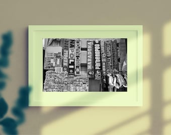May include: A black and white photograph in a light green frame showing a collection of sports scarves hanging on a wall. The scarves feature text including NAPOLI, SSC NAPOLI, MARADONA, PARTENOPEO, and FORZA NAPOLI. Below the scarves are small items.