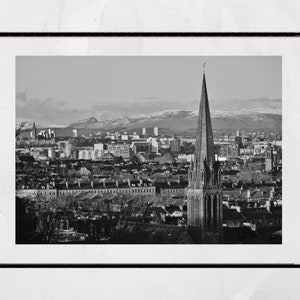 Könnte beinhalten: Ein Schwarzweißfoto einer Stadtlandschaft mit einem hohen Kirchturm im Vordergrund. Die Stadt liegt eingebettet in einem Tal mit schneebedeckten Bergen in der Ferne.