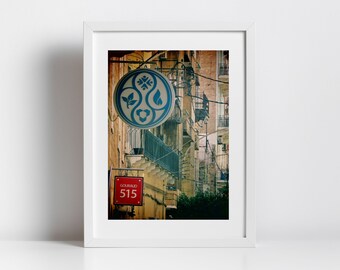 May include: A vintage photograph of a narrow street in a city. The building facades are a mix of yellow and beige with intricate details. A blue and white circular sign with a floral design hangs from the building. A red sign with the text "GOURAUD 515" is visible below the circular sign.