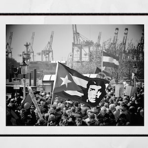 Op de afbeelding: Zwart-witfoto van een menigte mensen die vlaggen vasthouden, waaronder een Cubaanse vlag met een portret van Che Guevara. De menigte is verzameld voor een groot industriegebied met kranen en gebouwen.