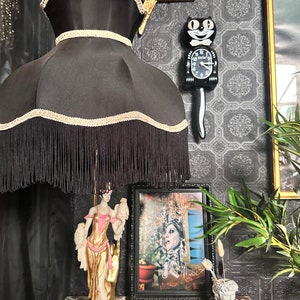 May include: A black lampshade with fringe and gold trim sits on a table lamp base with a gold and white figurine of a woman holding two birds. A black cat clock with a white face and gold hands is on the wall behind the lamp. A framed picture with a woman in a crown is on the table next to the lamp.