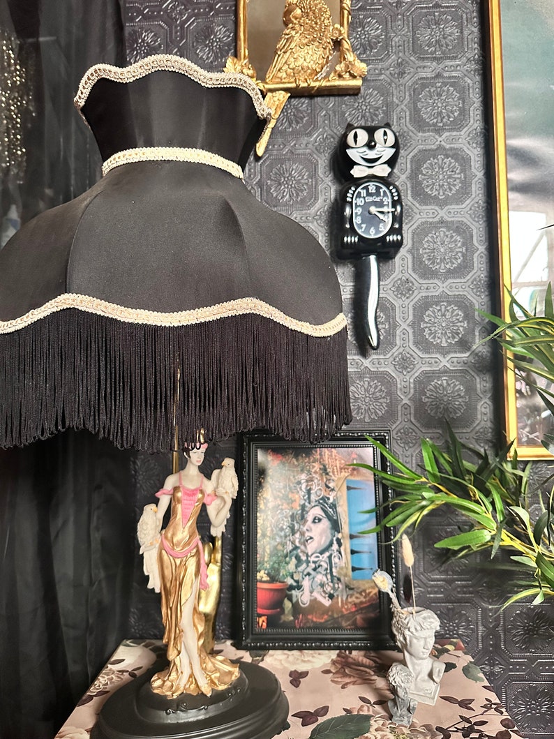 May include: A black lampshade with fringe and gold trim sits on a table lamp base with a gold and white figurine of a woman holding two birds. A black cat clock with a white face and gold hands is on the wall behind the lamp. A framed picture with a woman in a crown is on the table next to the lamp.