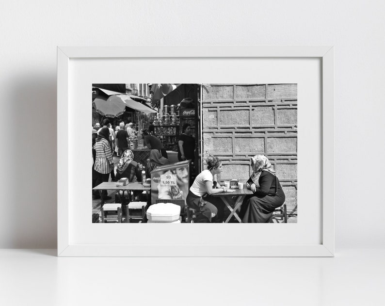 May include: Black and white photograph of a street scene with people. Two women sit at a small table, while others are in the background near a shop. The image is framed in a white border.