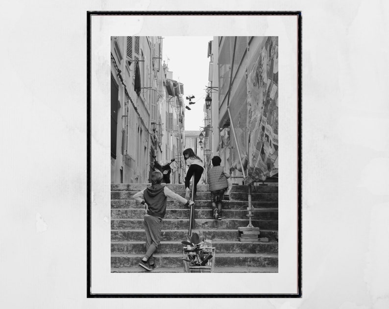 May include: Black and white framed photograph of children climbing a set of outdoor stairs in a narrow alleyway. One child leans on a metal railing, while two others ascend the steps. A shopping cart sits at the base of the stairs.