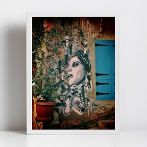 May include: A framed photograph of a street scene with a mural of a woman's face on a building. The mural is in black and white and the woman is wearing a crown. The building is painted in shades of orange, brown, and yellow. A blue door is visible on the right side of the image. A potted plant is in the foreground.
