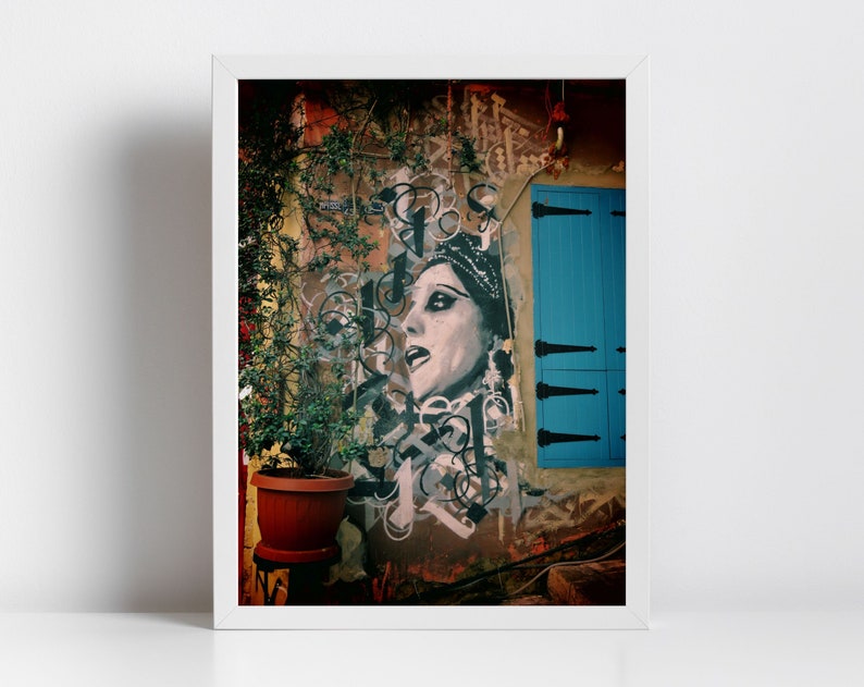 May include: A framed photograph of a street scene with a mural of a woman's face on a building. The mural is in black and white and the woman is wearing a crown. The building is painted in shades of orange, brown, and yellow. A blue door is visible on the right side of the image. A potted plant is in the foreground.