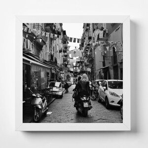 May include: A black and white photo of a narrow street in a city. A woman is riding a scooter down the street, with buildings and cars on either side. There are flags and laundry hanging from the buildings.