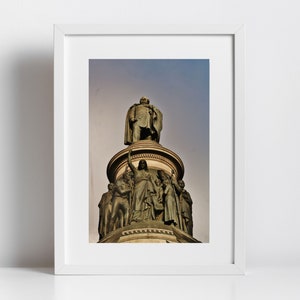 Puede incluir: Impresión enmarcada que muestra una estatua de bronce de un hombre con un abrigo largo sobre una columna. La estatua está rodeada de otras figuras. El cielo es una gradación de azul y gris.
