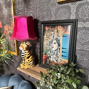 May include: A black framed picture with a colorful collage of a woman with a blue door in the background. The picture is on a shelf with a red lampshade, a tiger figurine, and a green plant.