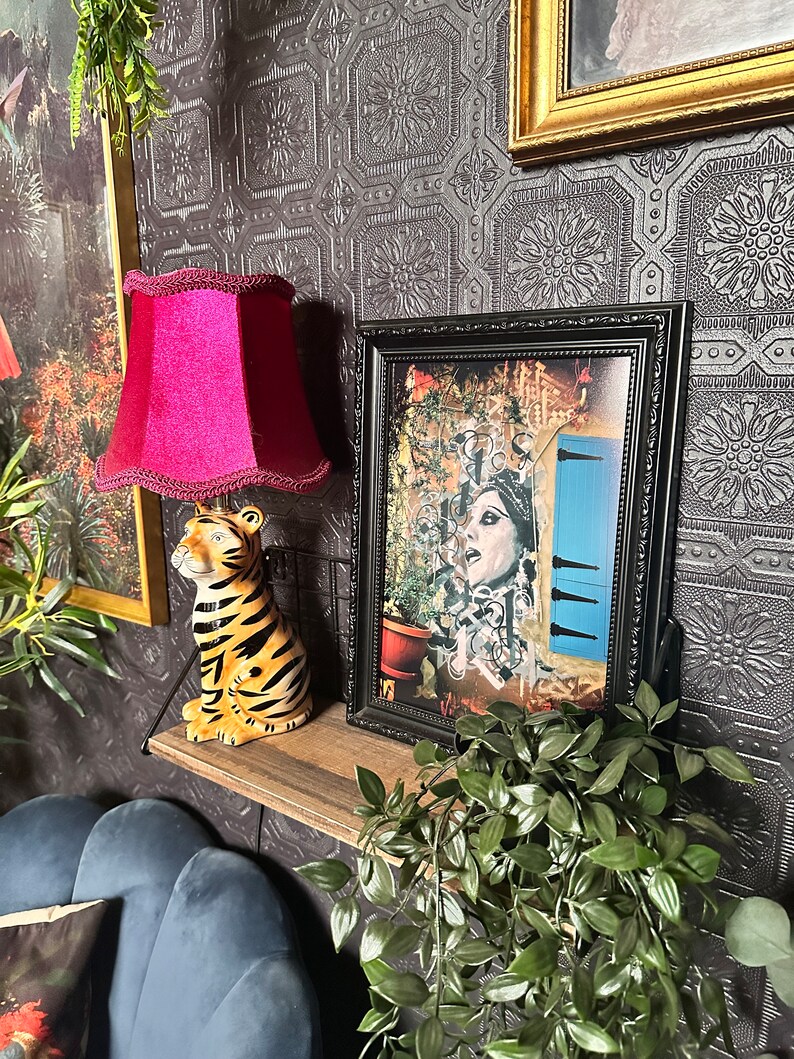 May include: A black framed picture with a colorful collage of a woman with a blue door in the background. The picture is on a shelf with a red lampshade, a tiger figurine, and a green plant.