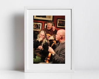 Könnte beinhalten: Gerahmtes Foto einer Gruppe von Musikern. Eine Frau mit langen grauen Haaren lacht, während sie einen Geigenbogen hält. Zwei Männer spielen Saiteninstrumente. Der Hintergrund zeigt gerahmte Bilder und eine rote Wand.